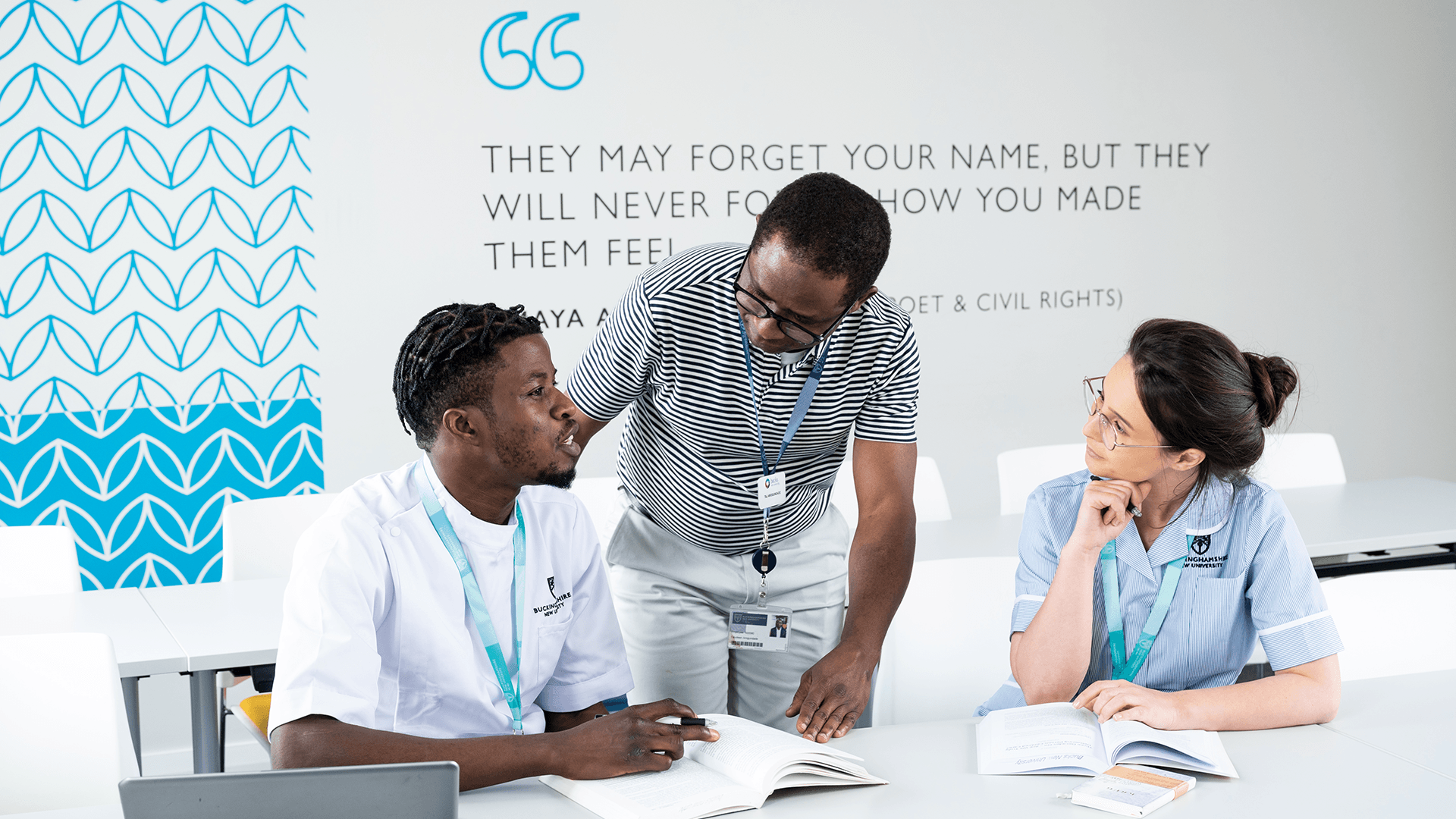 Two student nurses sat at a desk and a lecturer stood looking at the students talking through their work 