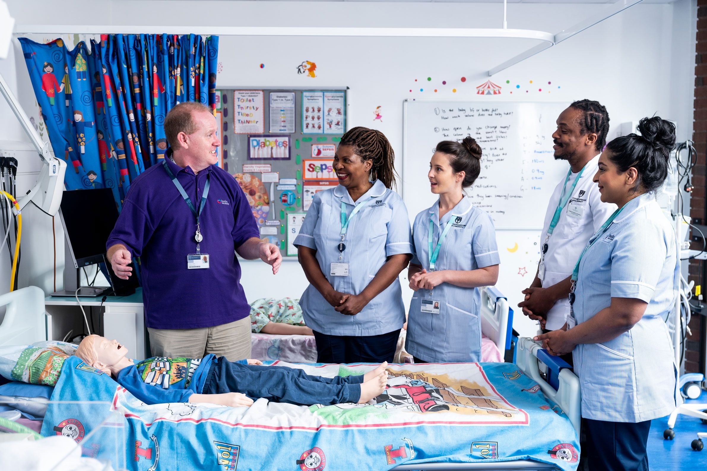 Four student nurses in uniform stood around a simulation hospital bed with a child laying down on it