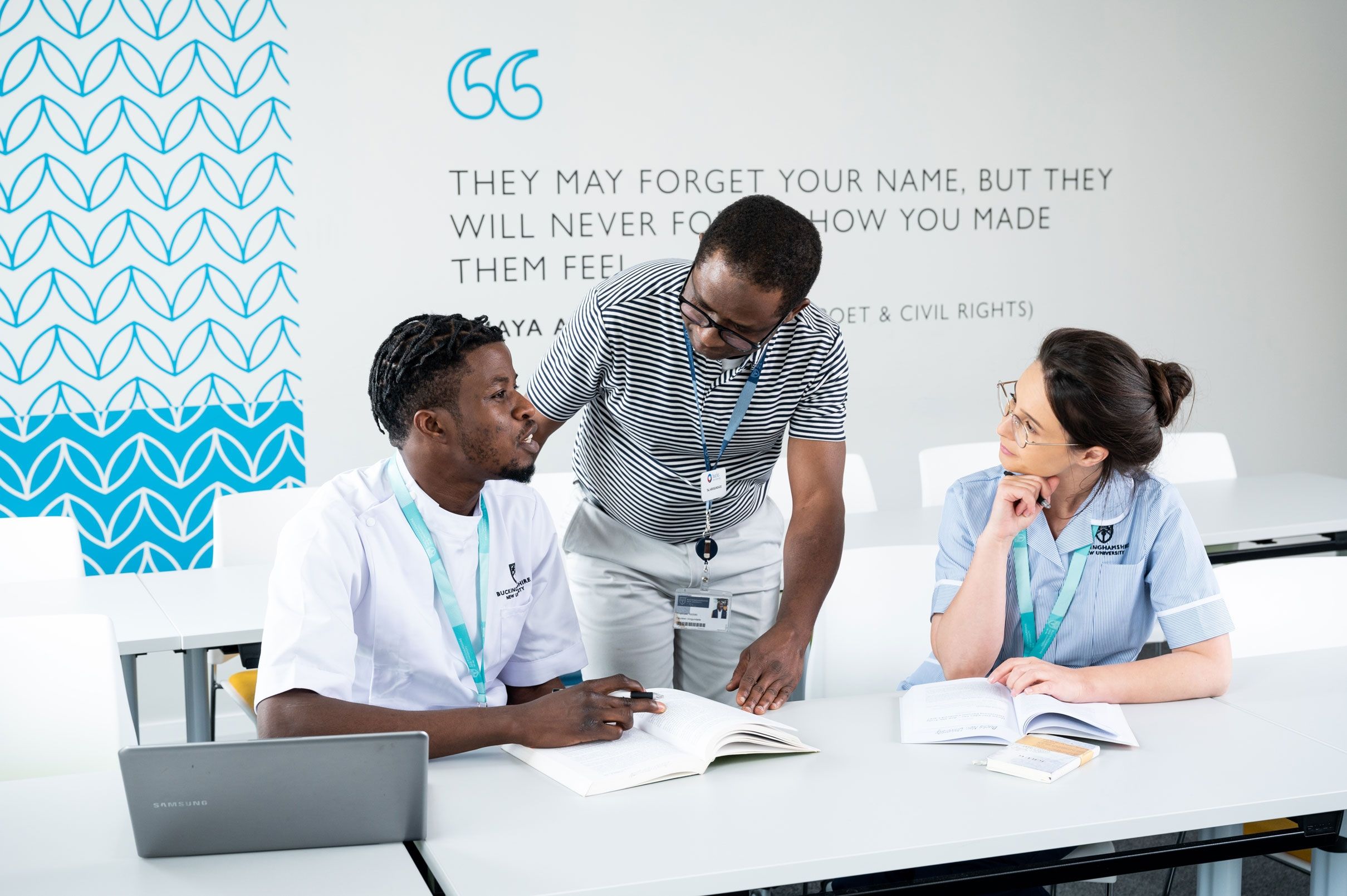 Two nursing students sat at a desk in a class with the teacher stood in between them in discussion