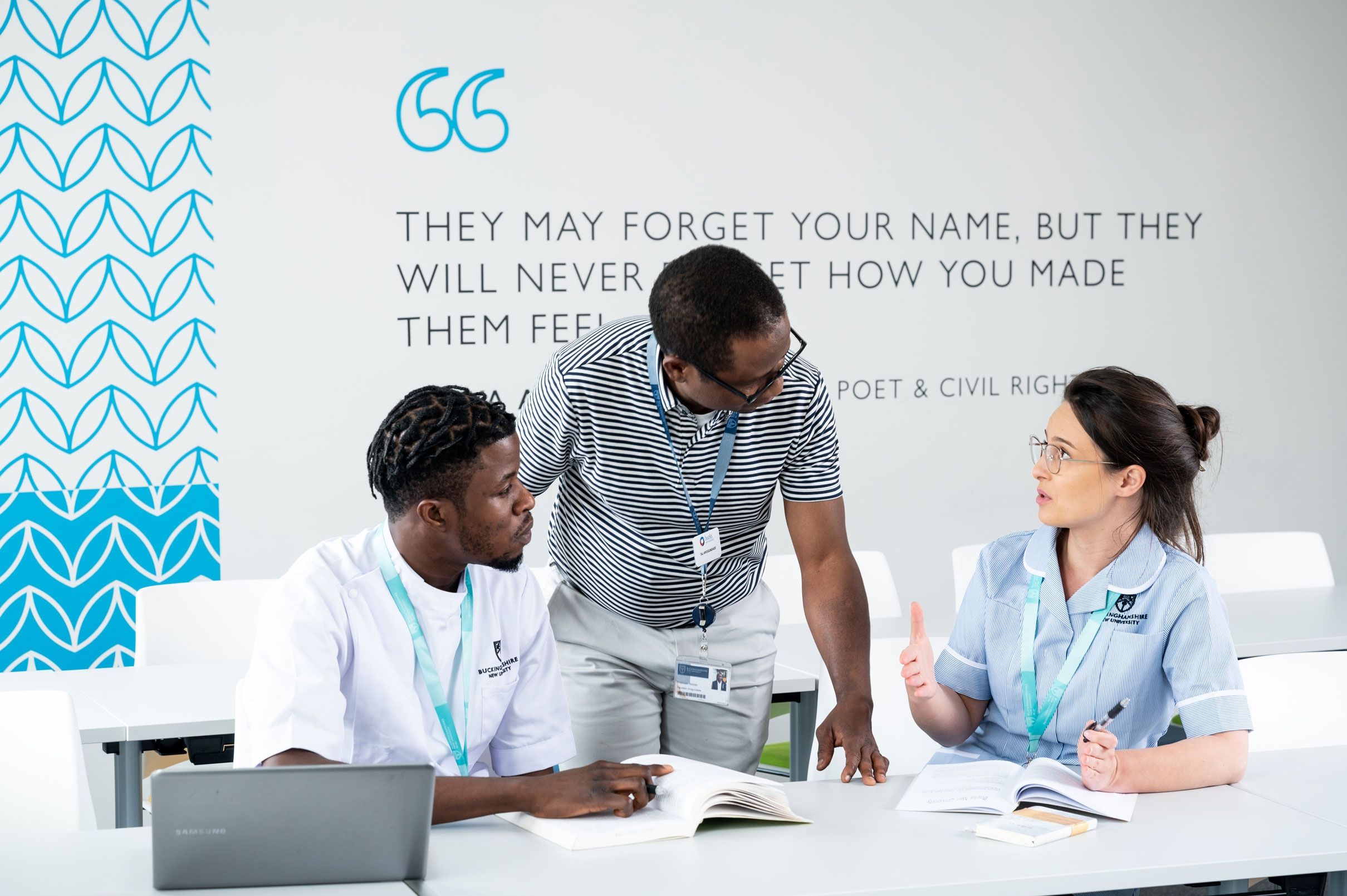 Two nursing students sat at a desk in a class with the teacher stood in between them in discussion