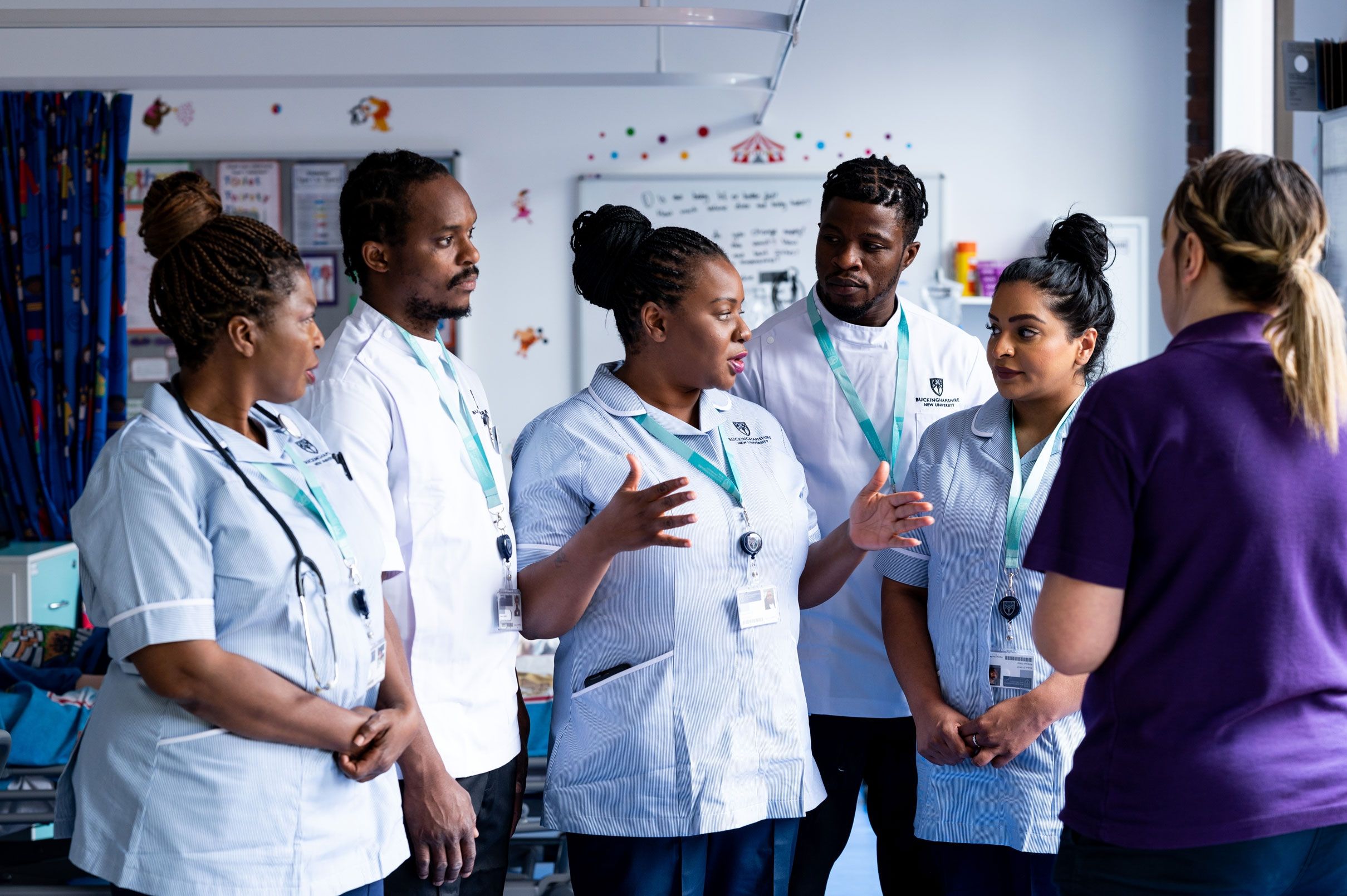 Five student nurses in uniform stood in discussion whilst facing a lecturer in a simulation ward