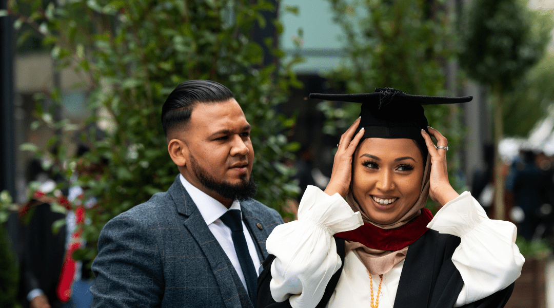 A graduate rearranging her hat whilst a man stood next to her looks at her in admiration