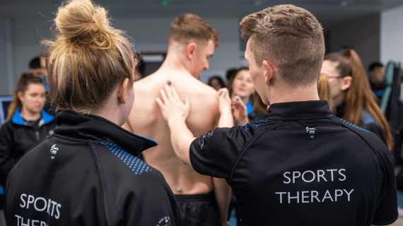 Two Sports Therapy students looking at the back of a fellow student, preparing for fitness testing. 