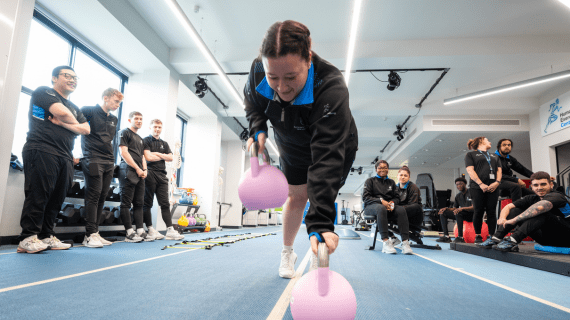 A Sports Therapy student on the indoor running track leaning down to pick up a weight from the floor.