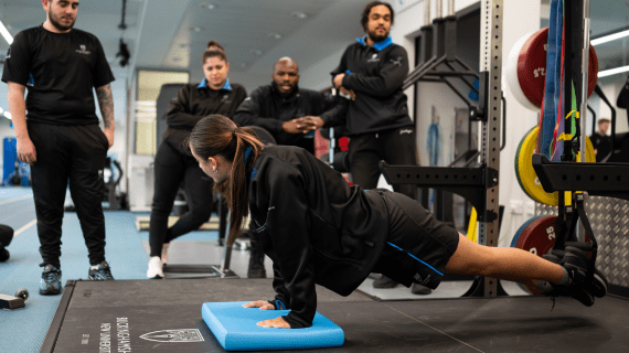 A Sports Therapy student doing a press up on the floor as four other students watch on.