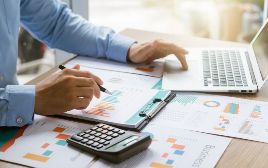 Someone sat at a desk with a laptop and calculator, whilst looking at graphs of data.