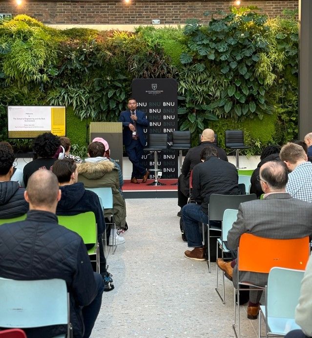 Room with people sat on seats and a speaker at the front holding a microphone and leaning on the podium