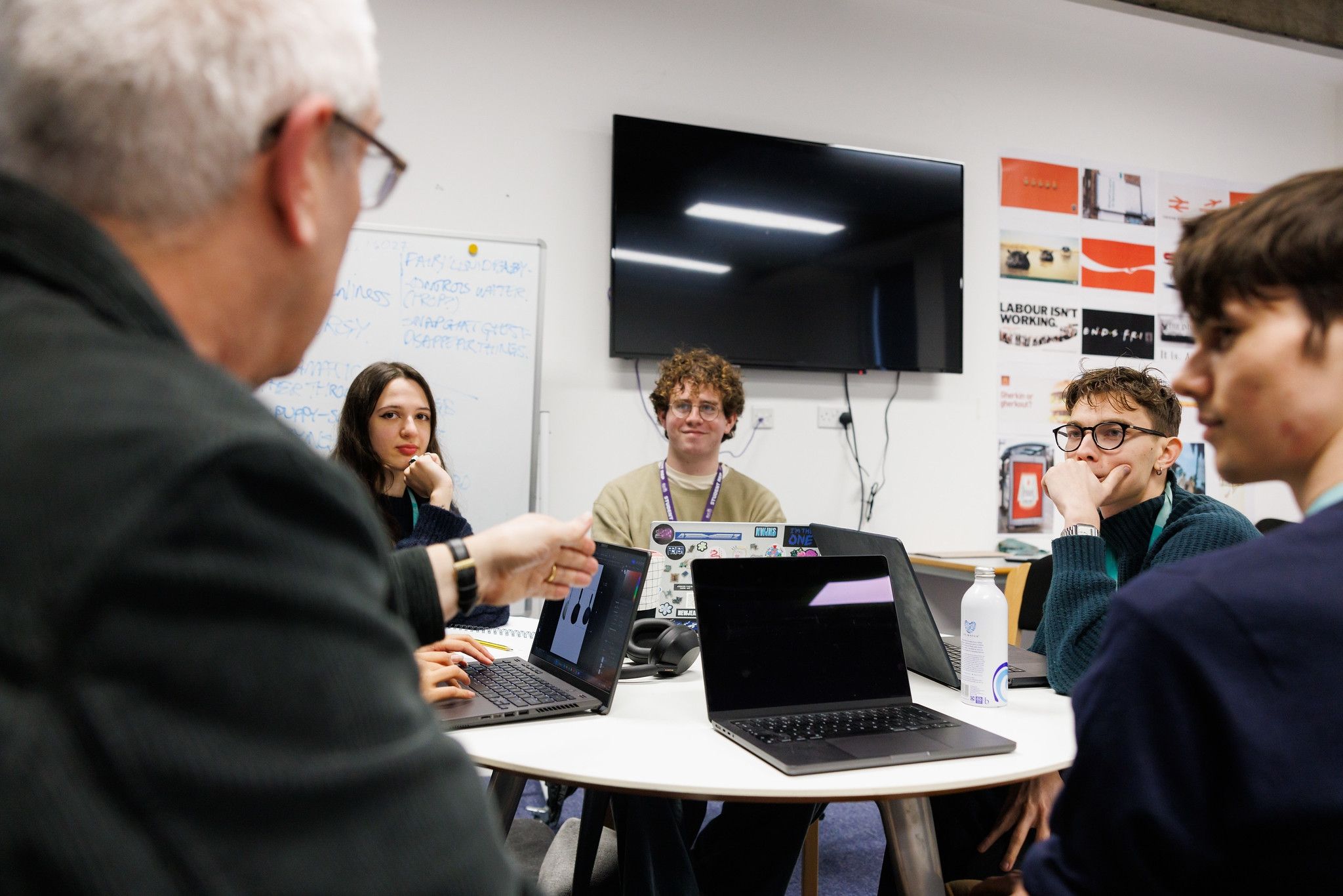 Students sitting around a table with their laptops, talking to a lecturer
