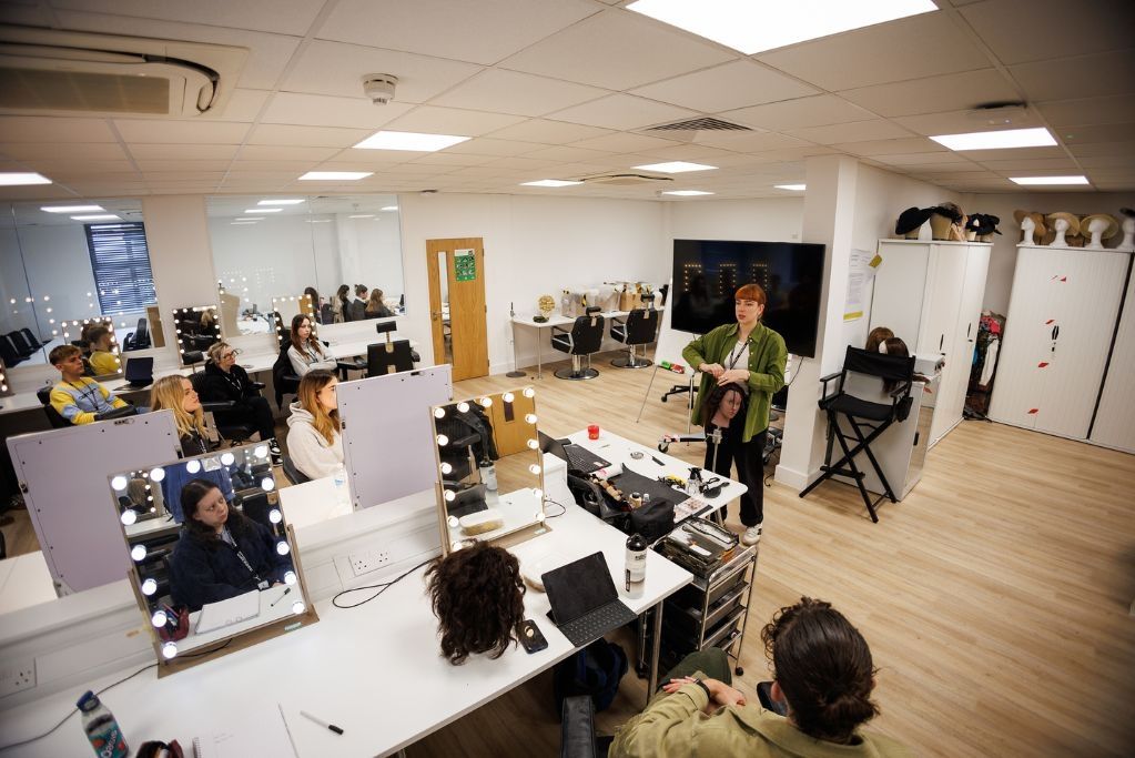 A Hair and Make-up lecturer stood at the front of a class holding a wig in front of students sat down at desks.