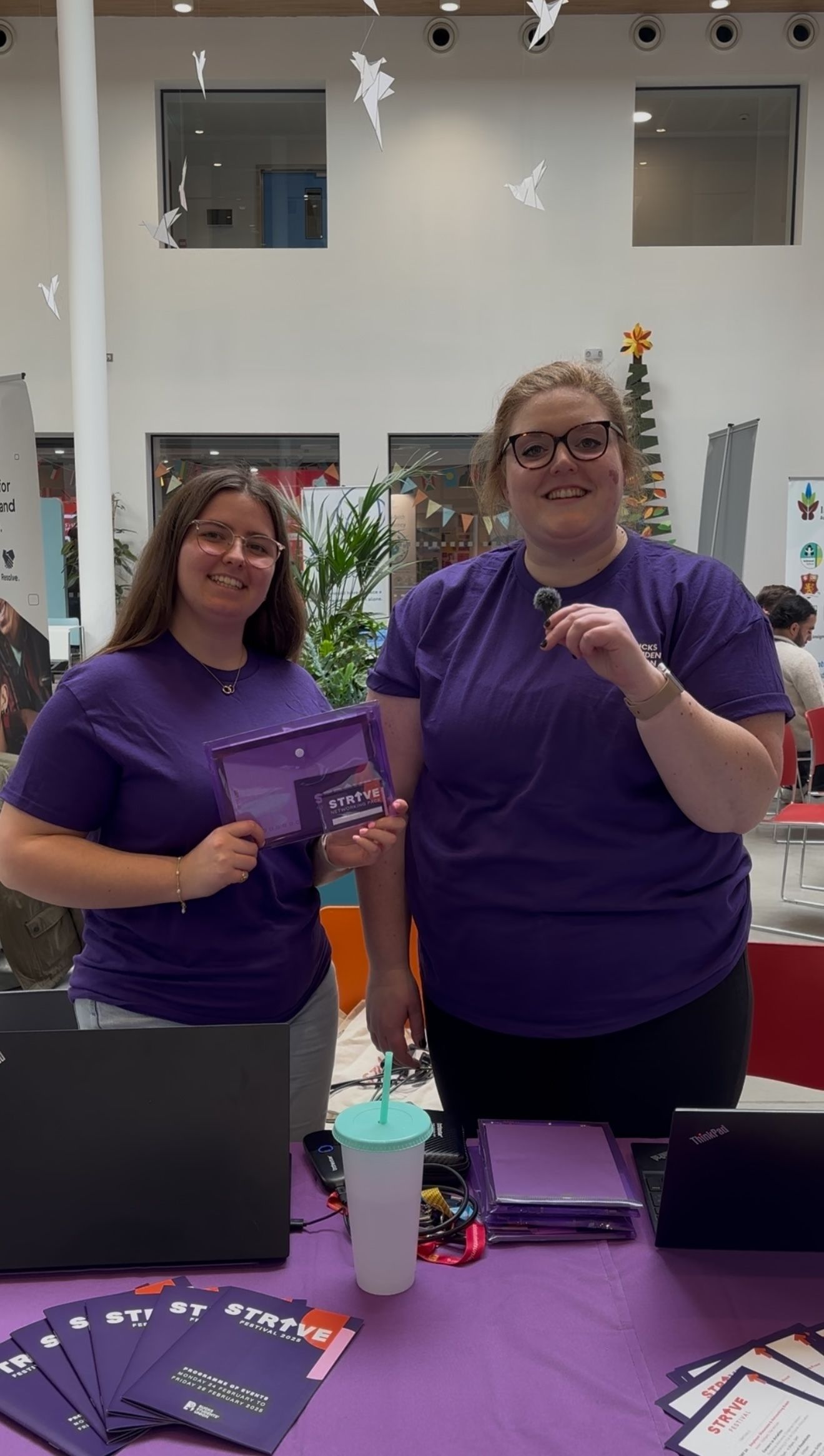 A picture of two women holding something in their hands and smiling at the camera. They are both in purple, the colour of the festival.