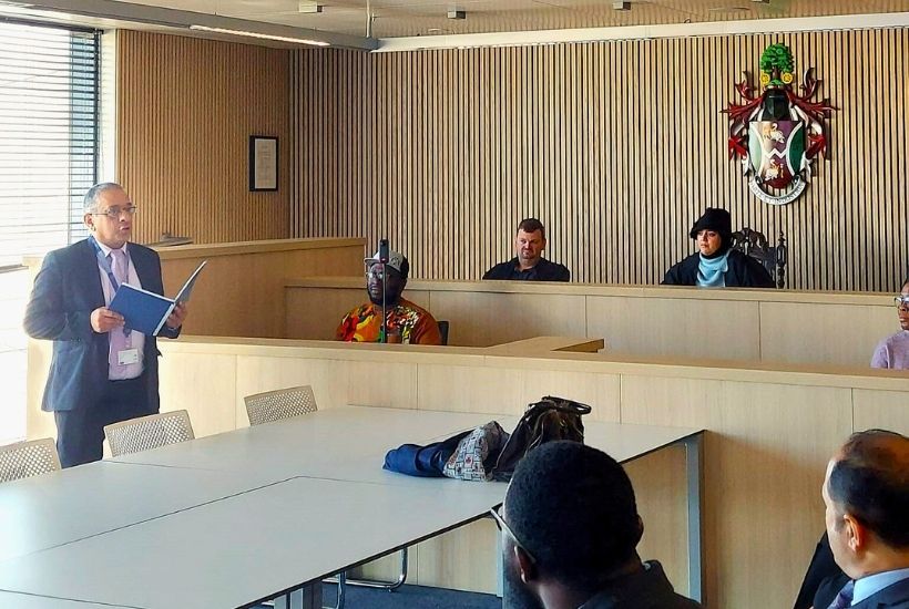 A man in a suit reads from a blue folder during a mock trial in a modern courtroom. Participants, a judge in robes, and several observers seated around a table.