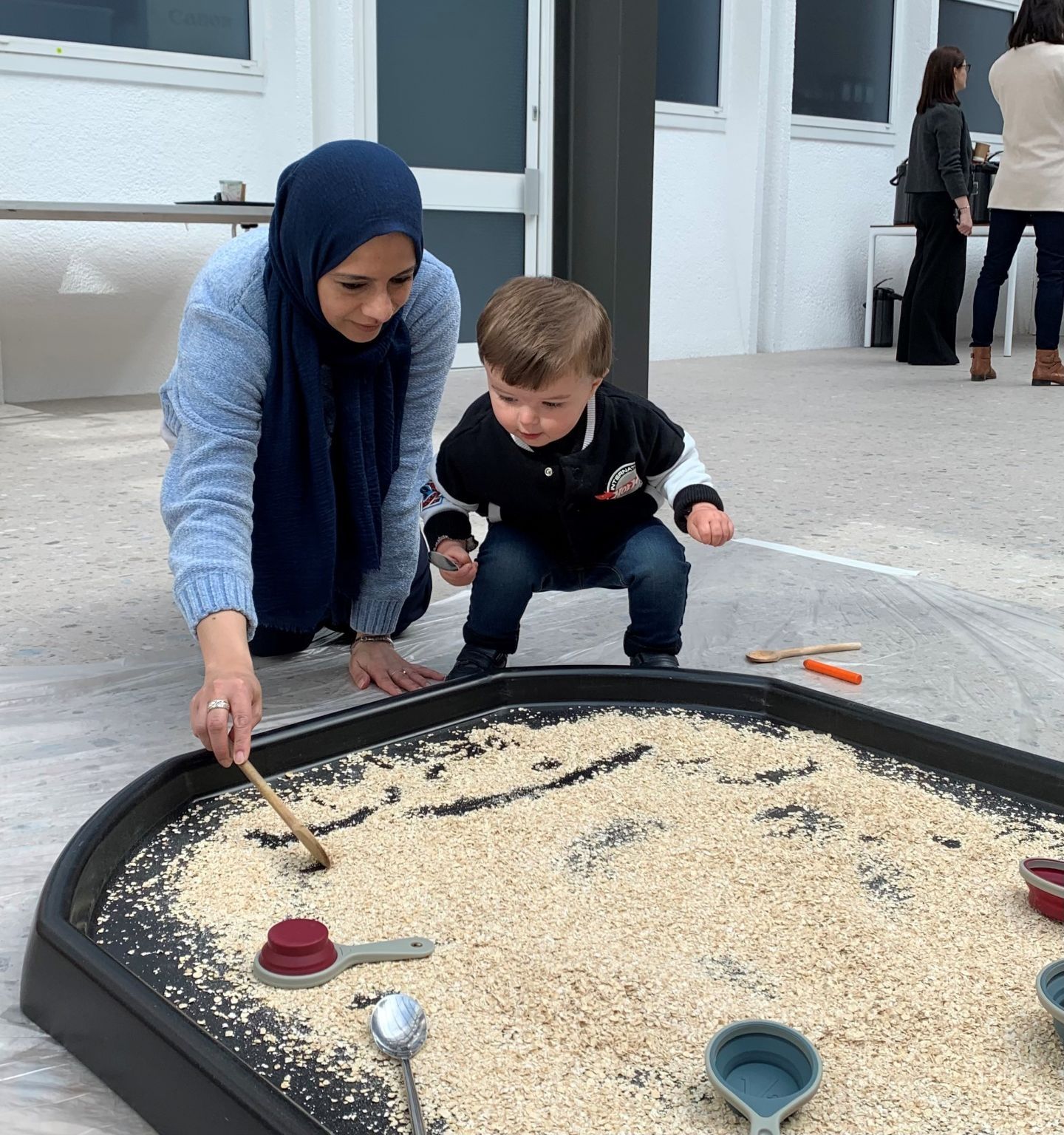 A woman plays with her child in front of sand pit.