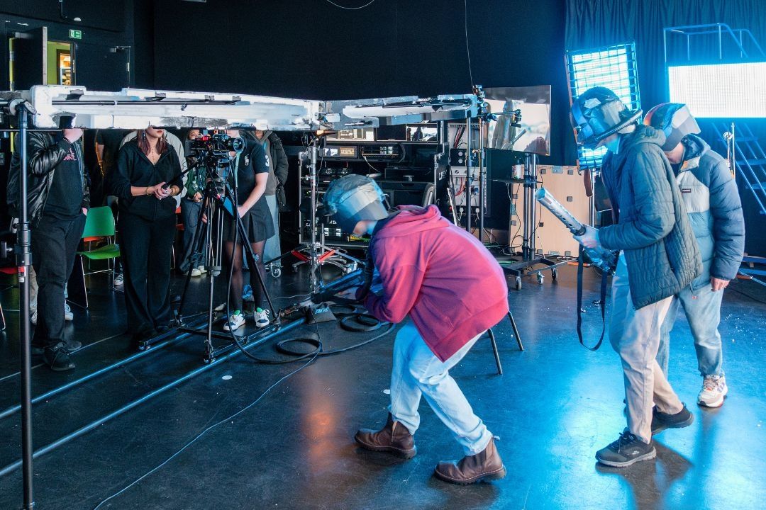 Three students acting on set whilst a group of Film students control the lighting and cameras behind scene.