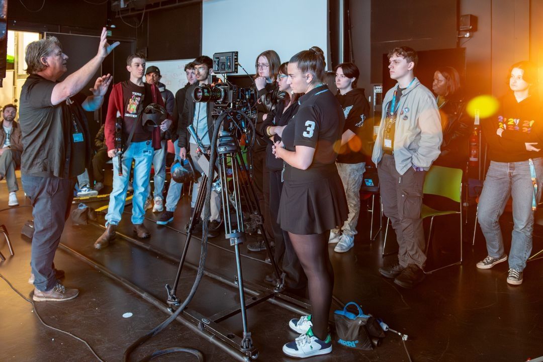 A group of students stood around a lecturer who has their hand raised whilst talking to the group in a Film and TV studio.