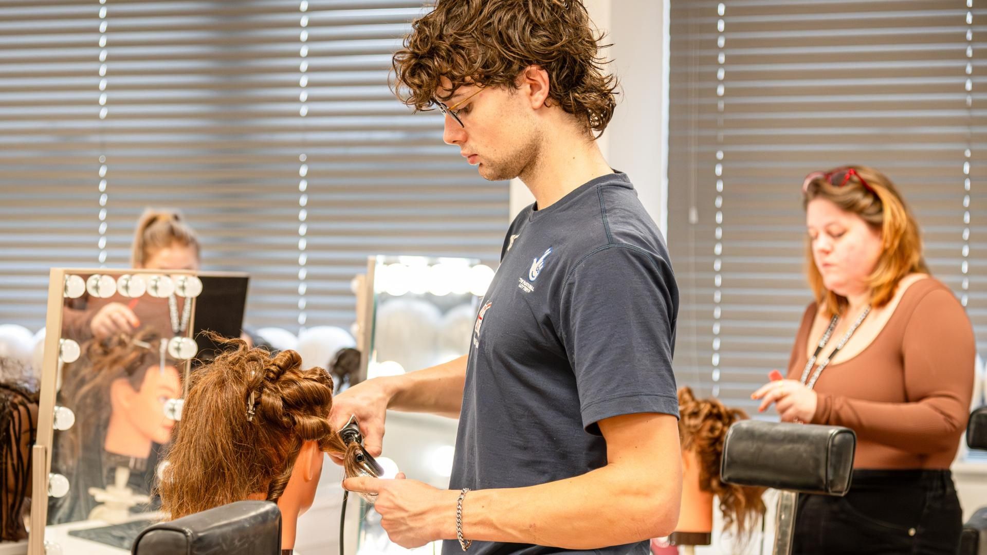 A Hair and make-up student stood up working on a wig in a classroom, with a fellow student in the back of the room..
