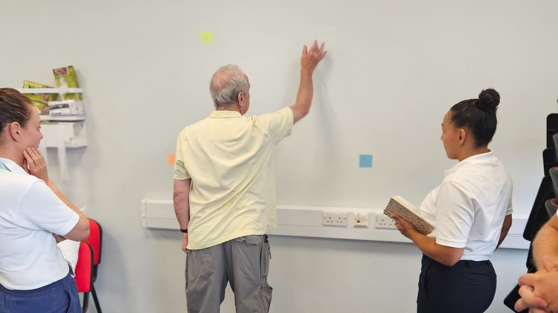 An elderly man raises his right hand against a wall as two Physiotherapy students watch on.