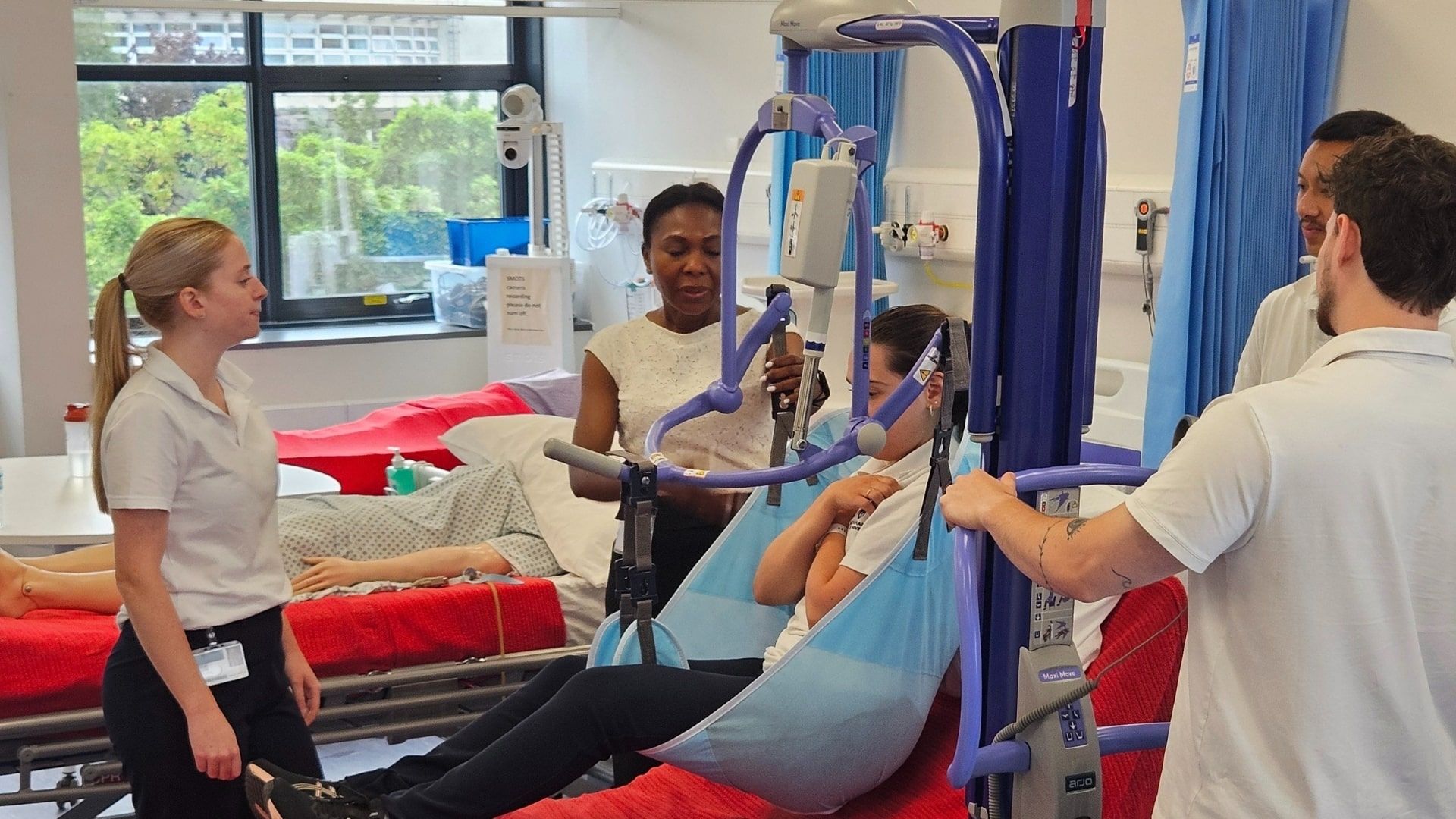 Three Physiotherapy students stood around a hospital simulation bed as a fellow student is sat in a hoist above a bed.