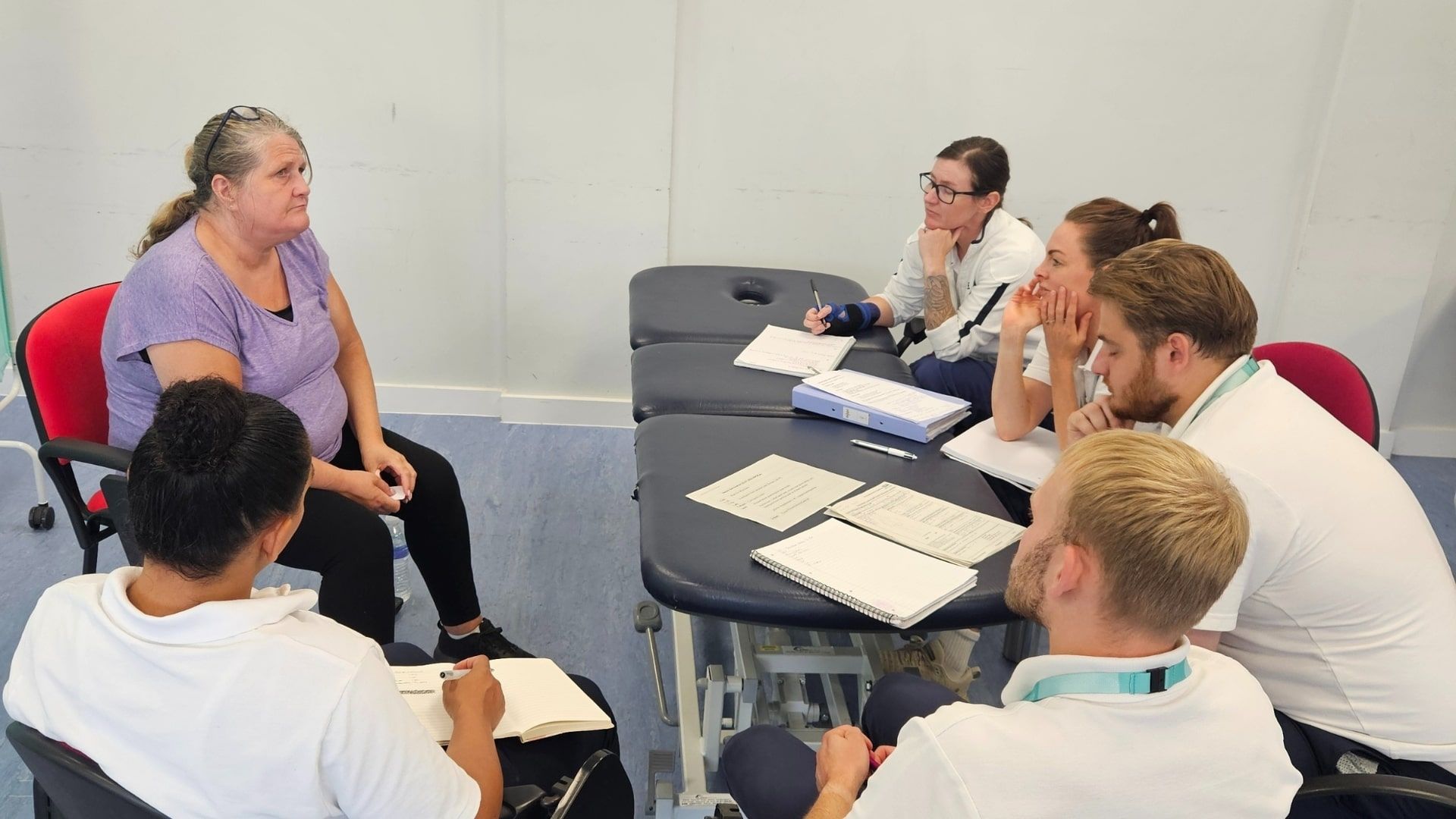 A member of the public sat on a chair in front of five Physiotherapy students with notebooks laid on a table.