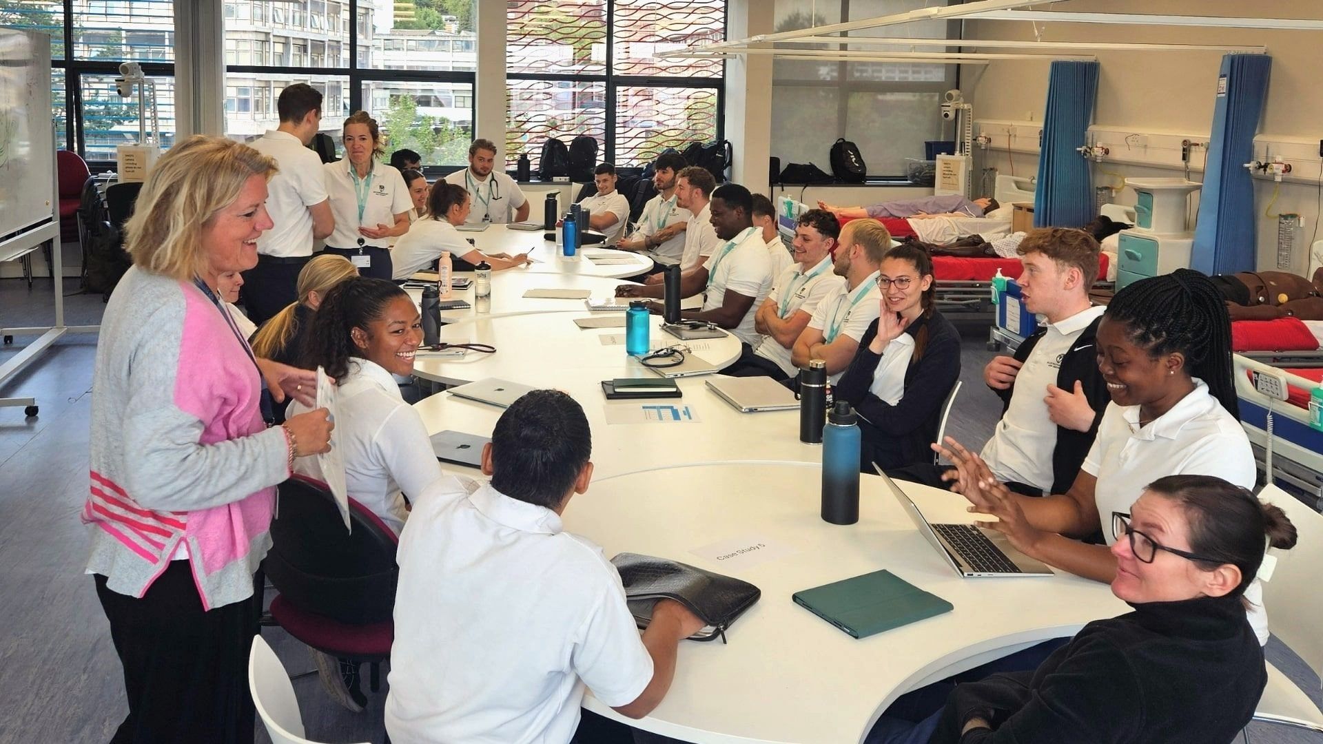 A class of Physiotherapy students sat round desks as a lecturer is stood on the left talking to students.