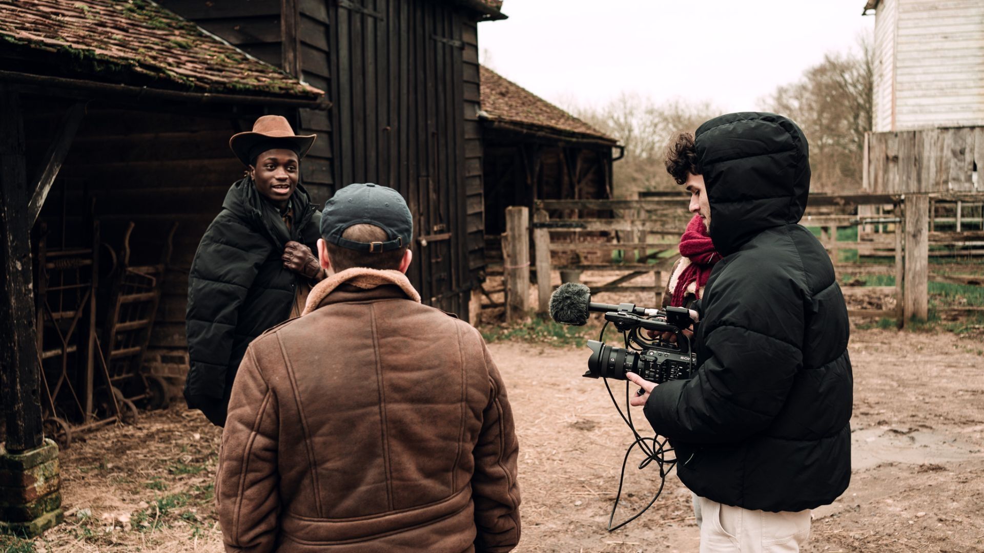 A cameraman wearing a coat stood next to another student as they both face a man wearing a cowboy cap in a stables setting.