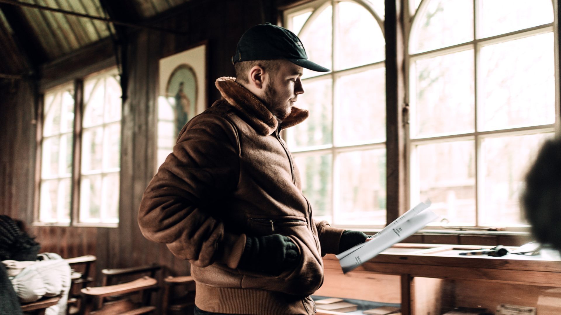 A student stood in an old buliding wearing a cap and holding a script in their left hand.