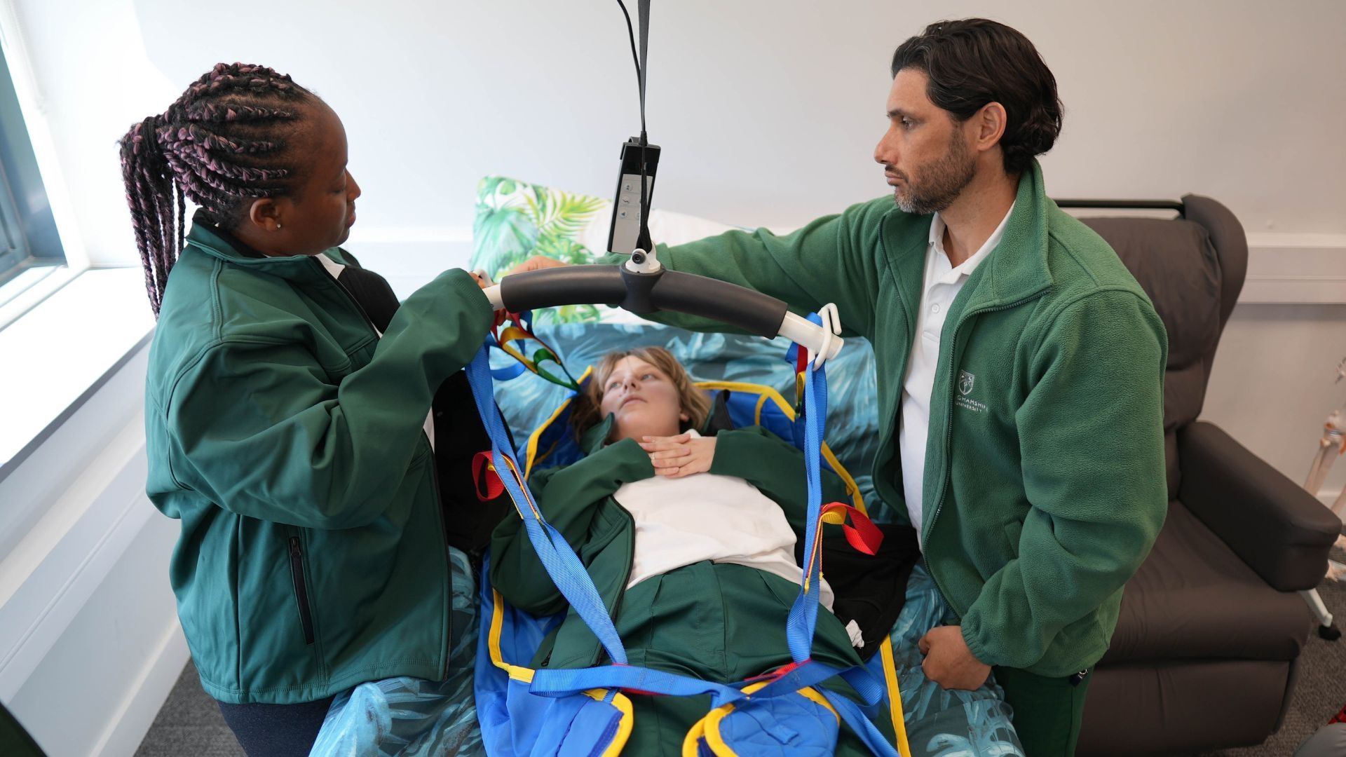 Occupational Therapy student in a hoist surrounded by two fellow students stood up either side.