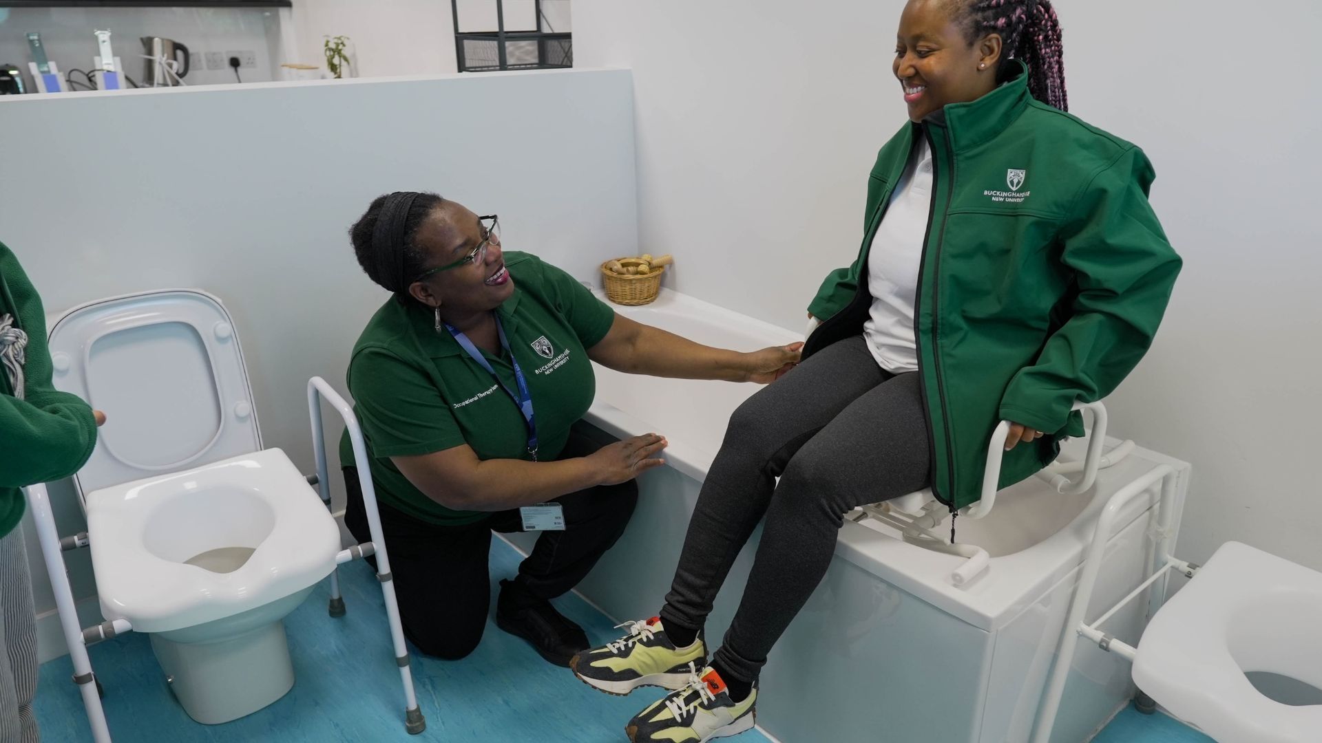 Two Occupational Therapy students in a bathroom. One sat on a seat in the bath and the other kneeling on the floor. 