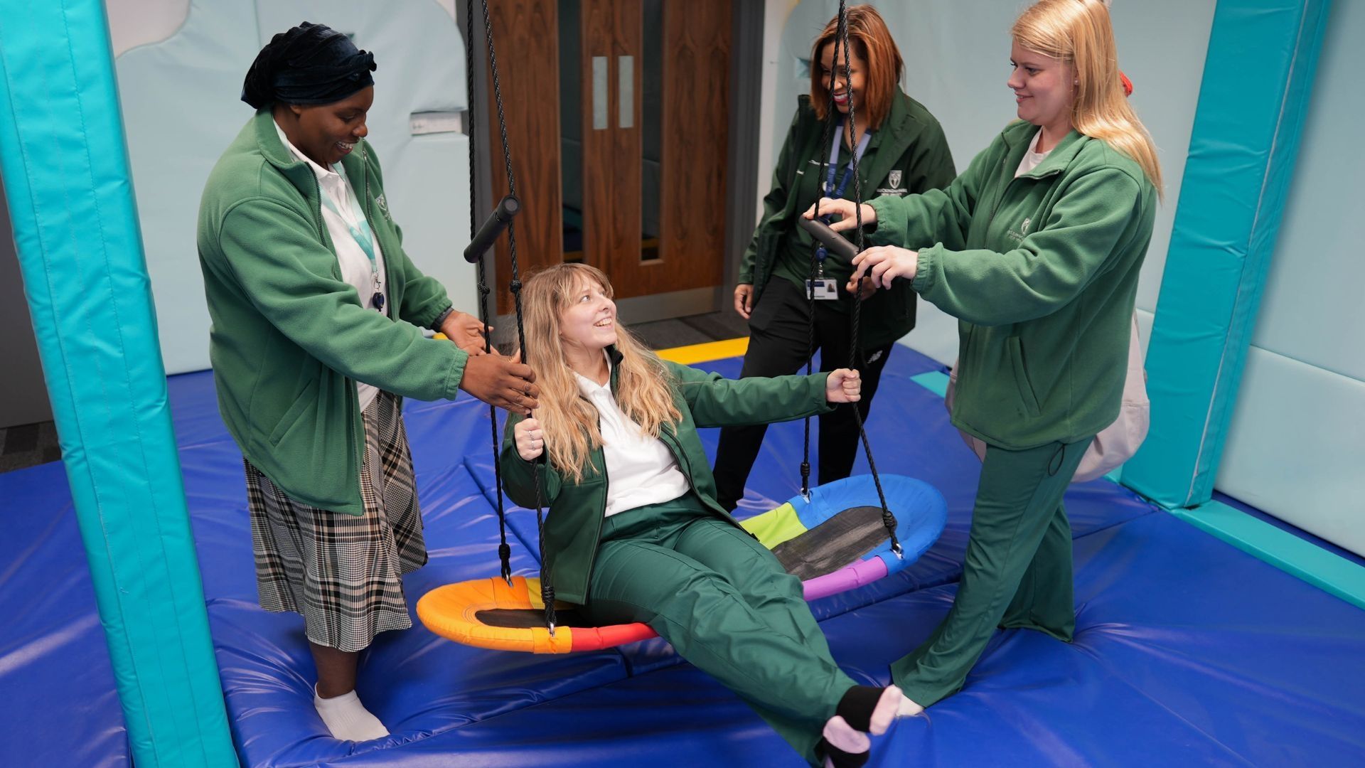 Three Occupational Therapy students stood either side of an indoor swing as another student holds onto the handles and swings.