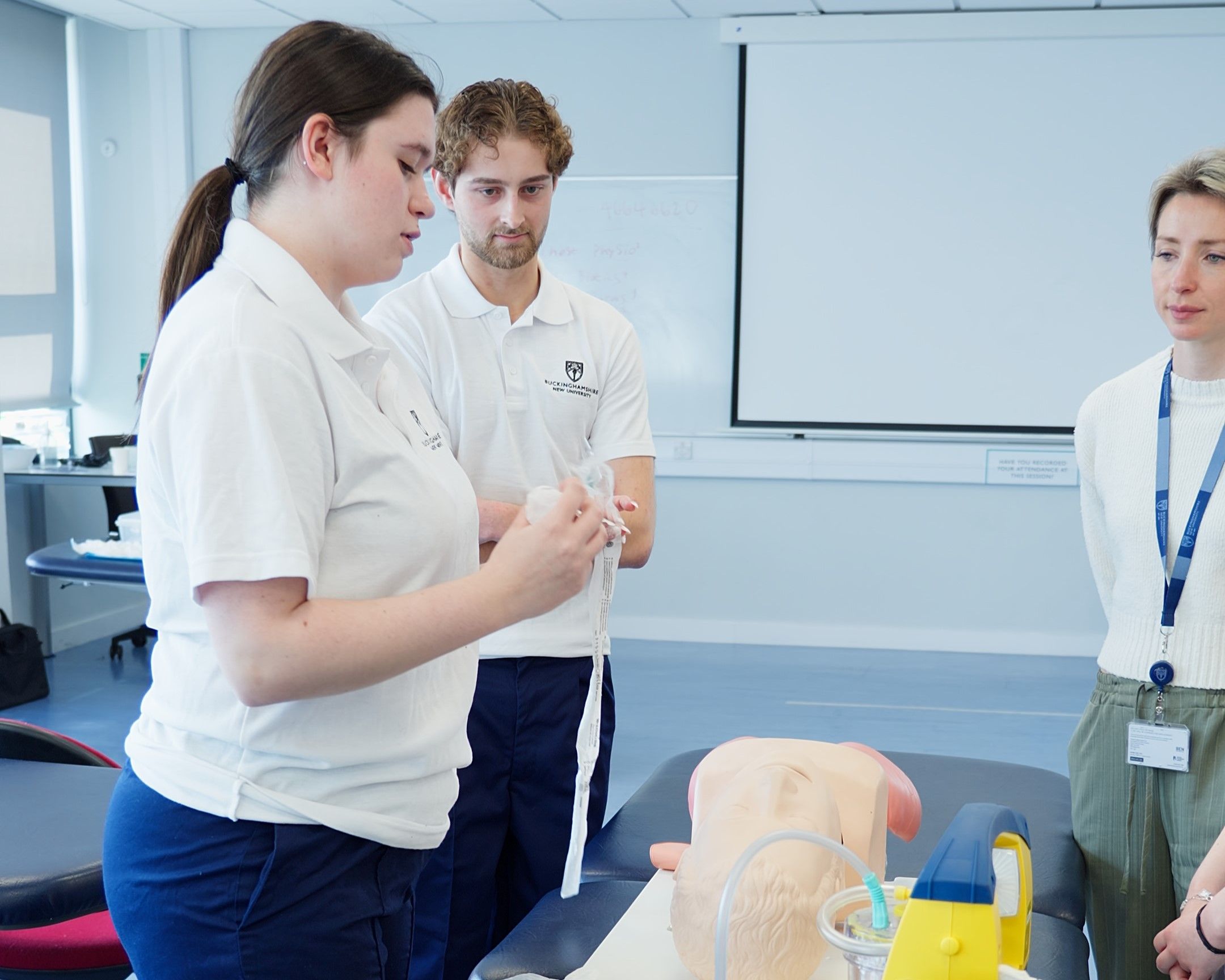 Physiotherapy students in white polo shirts practicing using medical suction equipment on a simulator mannequin during a practical session
