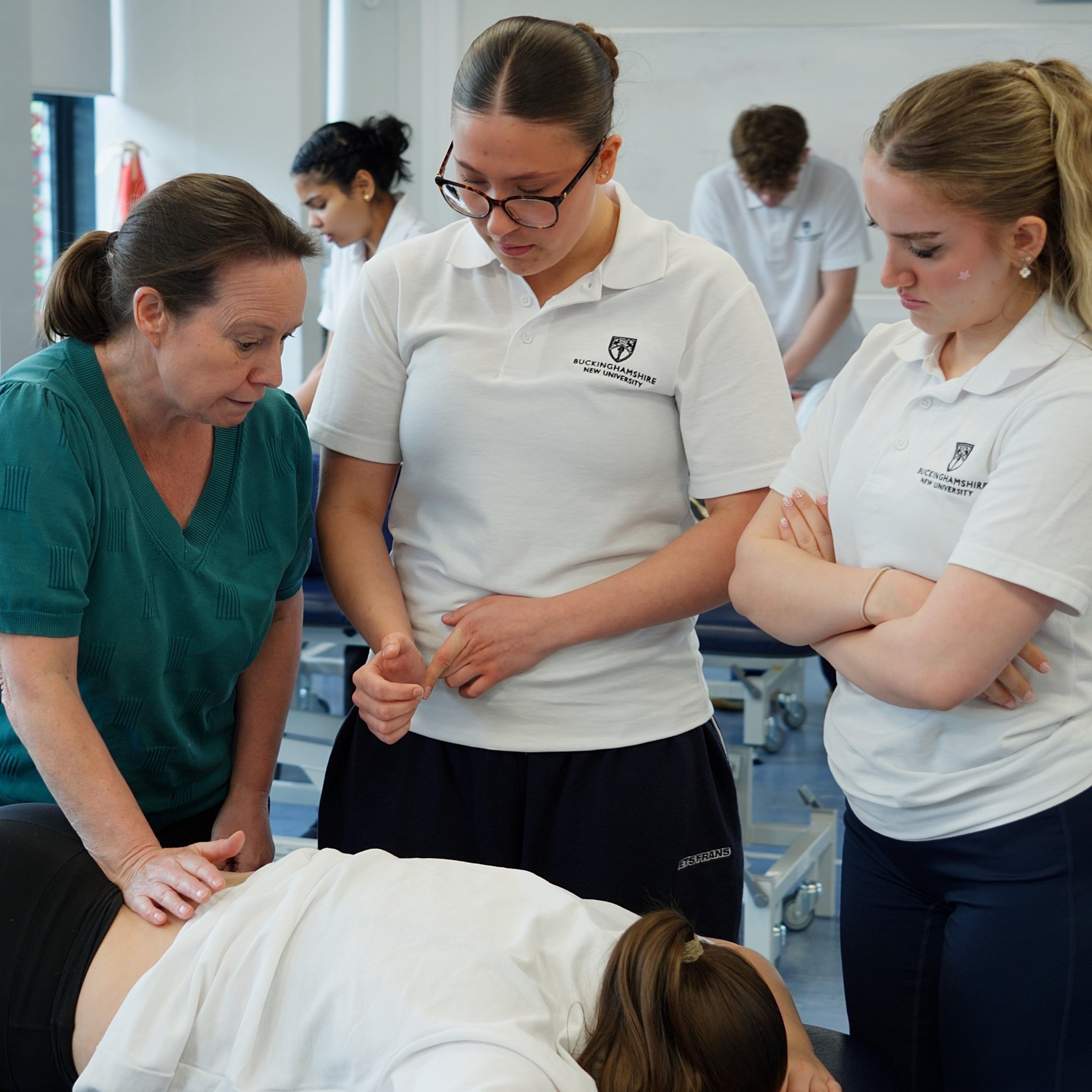 Two Physiotherapy students stood up observing a lecturer giving a massage on the lower back of a fellow student in a classroom setting.