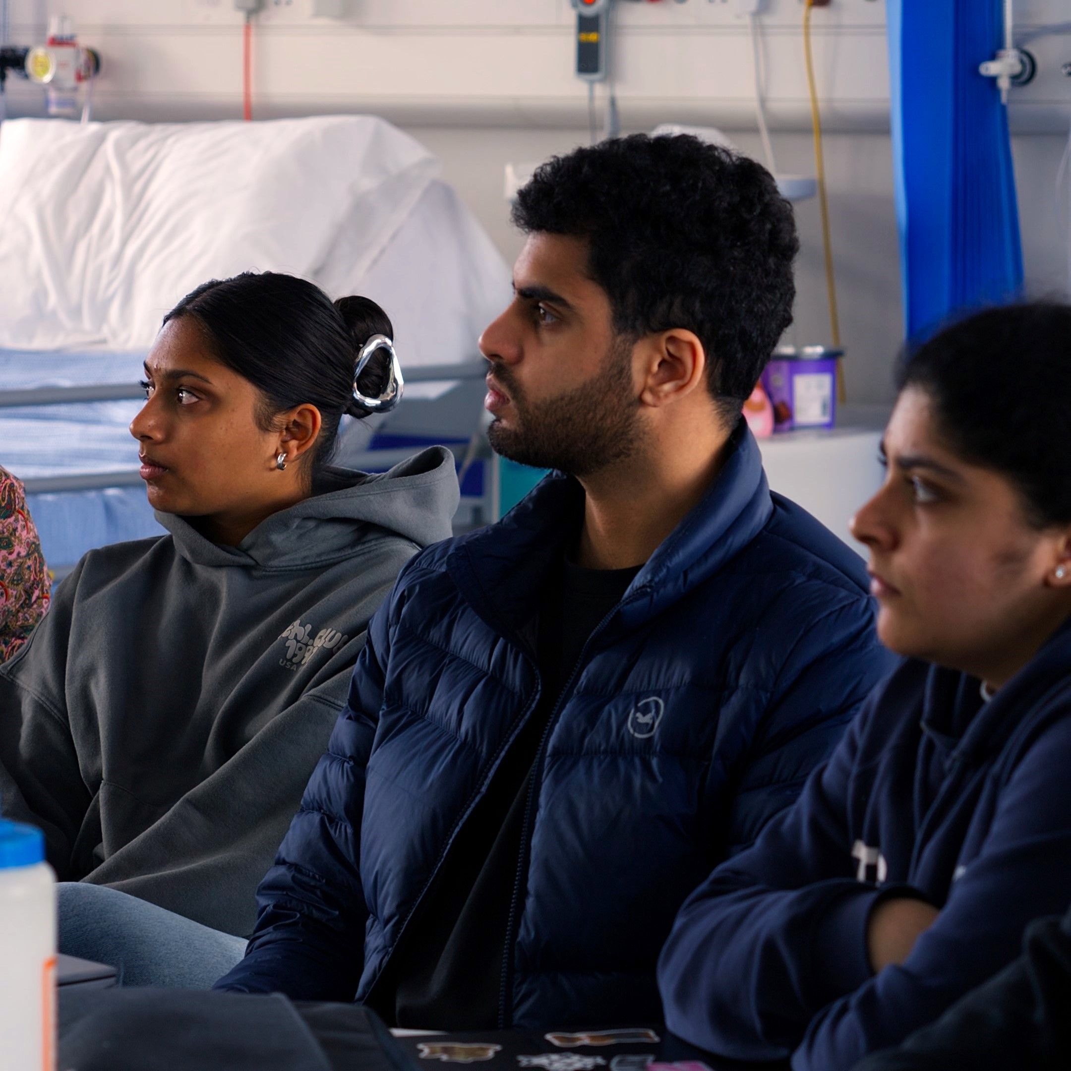 Three Physiotherapy students sat next to one another in a fake hospital setting with a concentrated look on their faces.